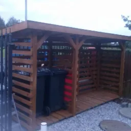 Drewniana konstrukcja osłaniająca dwa czarne pojemniki na śmieci, z ażurowymi ścianami z desek i podłogą z desek, na tle szarej żwirowej nawierzchni i ogrodzenia.