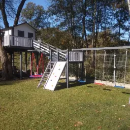 Kompleksowy plac zabaw z domkiem na drzewie, ścianką wspinaczkową i huśtawkami, pomalowany na szaro-biało, umieszczony na trawniku w ogrodzie.