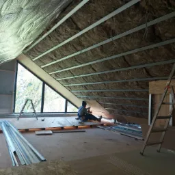 Prace wykończeniowe poddasza: robotnik w kombinezonie montuje płytę drewnianą na ocieplonym wełną mineralną stropie, widoczne profile metalowe, okna dachowe i drabina aluminiowa.