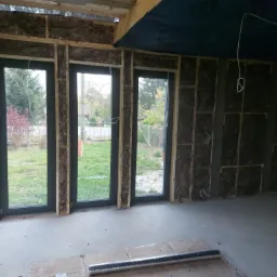 Wnętrze domu w trakcie prac, widoczne ocieplone ściany wełną mineralną, trzy okna z czarnymi ramami, nieotynkowana ściana z cegły po prawej, oraz materiały budowlane na podłodze.