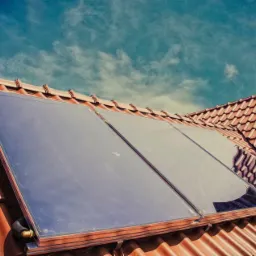 Panel kolektora słonecznego na dachu z ceglastą dachówką, odbijający błękitne niebo z chmurami. Widok z dołu, ukośny kadr.