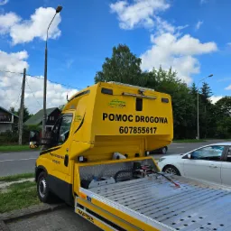 Żółta laweta z napisem 'Pomoc Drogowa' i numerem telefonu, zaparkowana przy ulicy, z widoczną platformą załadunkową i częściowo białym samochodem obok.