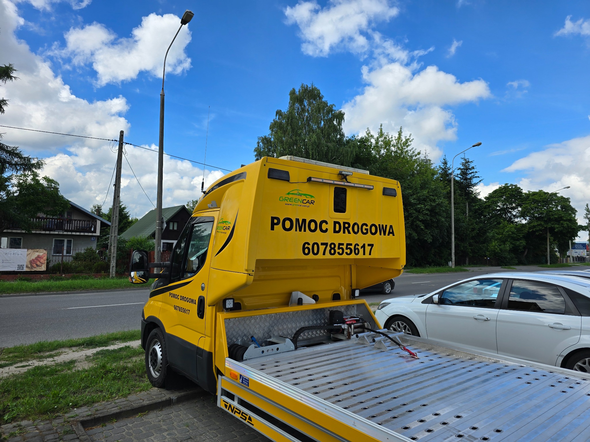 Żółta laweta z napisem 'Pomoc Drogowa' i numerem telefonu, zaparkowana przy ulicy, z widoczną platformą załadunkową i częściowo białym samochodem obok.