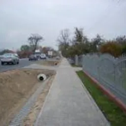 Nowy chodnik z kostki brukowej wzdłuż ulicy, z widoczną studzienką kanalizacyjną i metalowym ogrodzeniem. Tło: ruch uliczny i drzewa pod pochmurnym niebem.