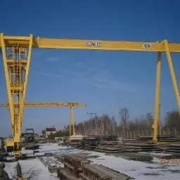 Żółty suwnicowy dźwig bramowy na placu budowy, podnosi betonowe płyty. Widoczny drugi dźwig w tle, błękitne niebo i częściowo ośnieżony teren.