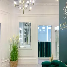 Eleganckie wnętrze salonu kosmetycznego z marmurkową posadzką, złotym żyrandolem i zieloną sofą z weluru. W tle widoczna recepcja z zielonymi panelami.