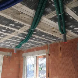 Niebieskie i zielone rury karbowane przymocowane do sufitu w surowym stanie, widoczne ściany z czerwonej cegły i okna.