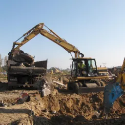 Żółta koparka CAT M315 załadowuje gruz na ciężarówkę na placu budowy. Widoczny pracownik w kamizelce. W tle drzewa i błękitne niebo.