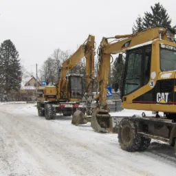 Dwa żółte, kołowe  Caterpillar M315 na zaśnieżonej drodze; w tle budynki i drzewa. Jeden z nich z przodu po prawej stronie kadru, drugi w oddali po lewej stronie.