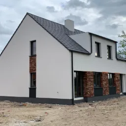 Dwukondygnacyjny dom z białym tynkiem i fragmentami elewacji z czerwonej cegły, szara dachówka, widoczne pozostałości po budowie