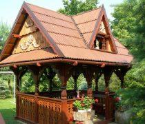 Drewniana altana ogrodowa z dekoracyjnymi rzeźbieniami, brązowy dach z dachówki, balustrada z doniczkami kwiatów, w otoczeniu zieleni.
