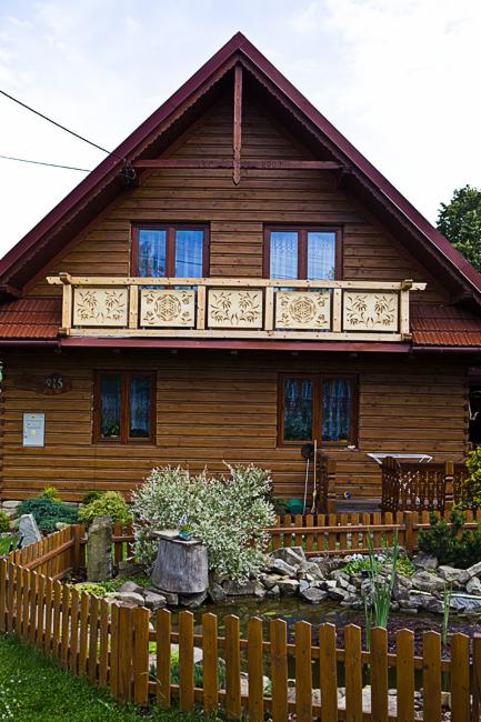 Drewniany dom z balkonem zdobionym ażurowymi panelami. Uwagę przykuwa mały staw ogrodowy z drewnianym płotkiem na pierwszym planie. Widoczne okna z brązowymi ramami.