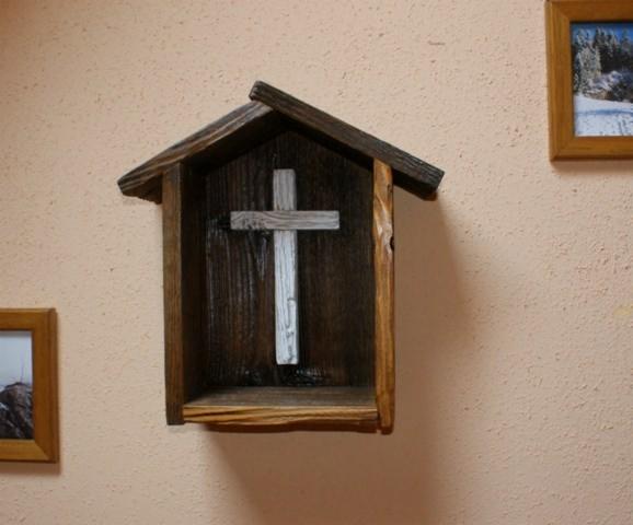 Drewniana, ręcznie robiona kapliczka w kształcie domku, z białym krzyżem w środku, zawieszona na teksturowanej ścianie w ciepłym odcieniu. Obok wiszą dwa obrazy w drewnianych ramach.