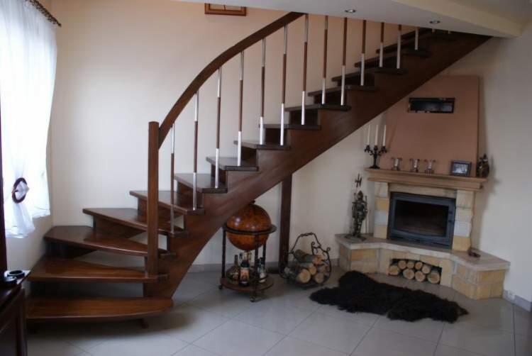 Kręte, drewniane schody z metalowymi poręczami prowadzą na górę, obok kominka z dekoracjami. Globus-barek dodaje elegancji wnętrzu.