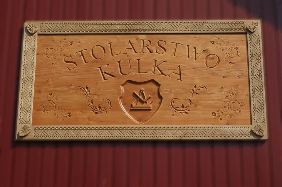 Ręcznie rzeźbiony, drewniany szyld z napisem 'Stolarstwo Kulka', zdobiony ornamentami i herbem, zawieszony na ciemnoczerwonej ścianie.