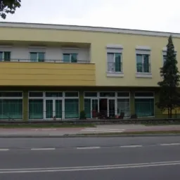 Żółty budynek z białymi oknami i roletami, widok z ulicy. Na parterze duże okna wystawowe. Architektura miejska, zieleń w tle, spokojna okolica.