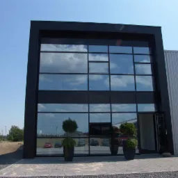 Nowoczesna fasada budynku z dużymi, aluminiowymi oknami, w których odbija się błękitne niebo. Przed wejściem ustawione dekoracyjne rośliny w donicach.