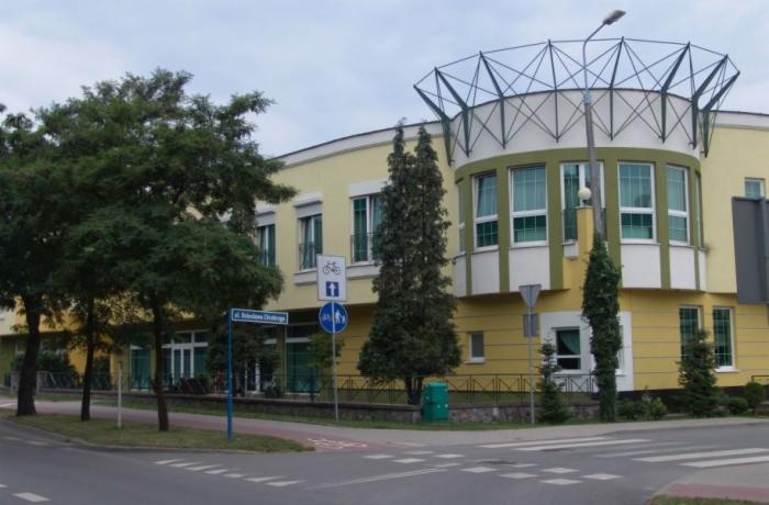 Żółty budynek z białymi oknami i zielonymi akcentami architektonicznymi, widziany z ulicy w pochmurny dzień. Widoczne drzewa i znaki drogowe.