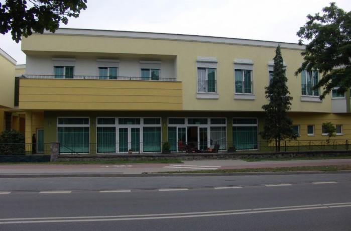 Żółty budynek z białymi oknami i roletami, widok z ulicy. Na parterze duże okna wystawowe. Architektura miejska, zieleń w tle, spokojna okolica.