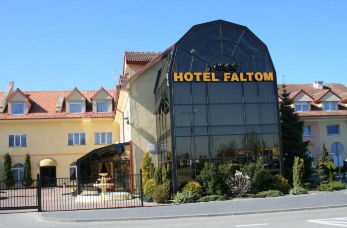 Elewacja hotelu Faltom z przeszkloną częścią, widoczne okna, dach z czerwonej dachówki, brama wjazdowa i zieleń. Budynek o żółtej elewacji w słoneczny dzień.