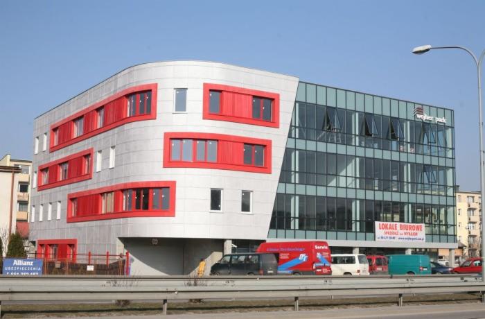 Nowoczesny budynek biurowy z aluminiową fasadą i czerwonymi obramowaniami okien, widoczny z poziomu ulicy w słoneczny dzień. Część budynku ma szklaną fasadę.