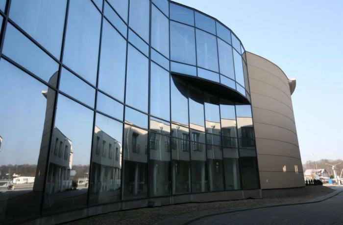 Nowoczesny budynek biurowy z fasadą ze szkła refleksyjnego, w której odbija się otoczenie. Architektura z zaokrąglonymi elementami i dużymi przeszkleniami.