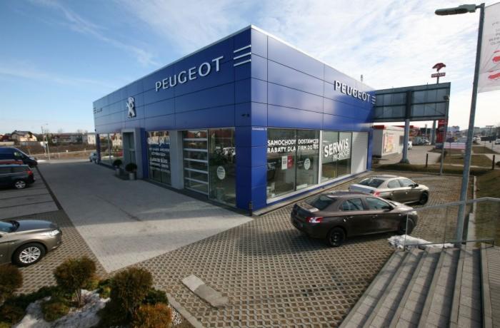 Nowoczesny budynek salonu samochodowego Peugeot z przeszklonymi drzwiami i oknami, na zewnątrz zaparkowane samochody. Błękitna elewacja z logo marki.