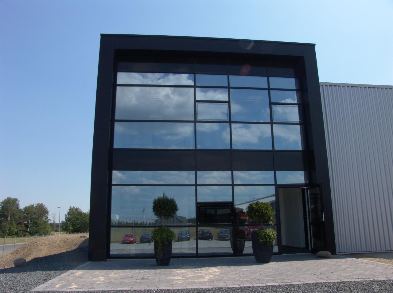 Nowoczesna fasada budynku z dużymi, aluminiowymi oknami, w których odbija się błękitne niebo. Przed wejściem ustawione dekoracyjne rośliny w donicach.