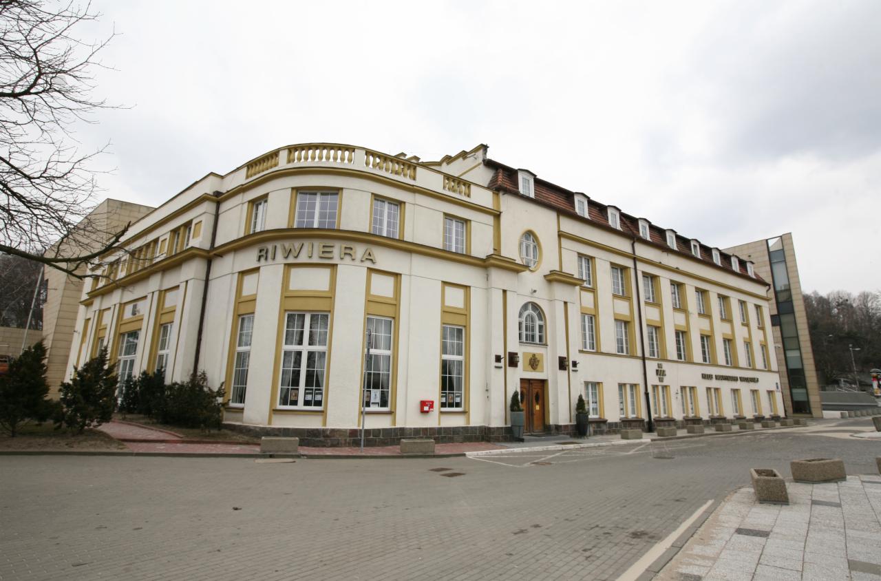 Elewacja budynku Riviera z jasną fasadą i żółtymi detalami architektonicznymi, widoczne liczne okna, wejście z drewnianymi drzwiami i otoczenie z kostki brukowej.