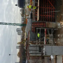 Wysoka konstrukcja żelbetowa w trakcie budowy, widoczne szalunki, rusztowania, zbrojenia i pracownicy w żółtych kamizelkach na tle miejskiego krajobrazu pod pochmurnym niebem.