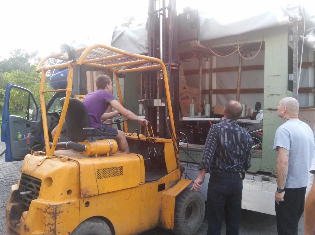 Żółty wózek widłowy z operatorem załadowuje maszynę do ciężarówki. Obserwatorzy w tle. Przewóz gabarytów.