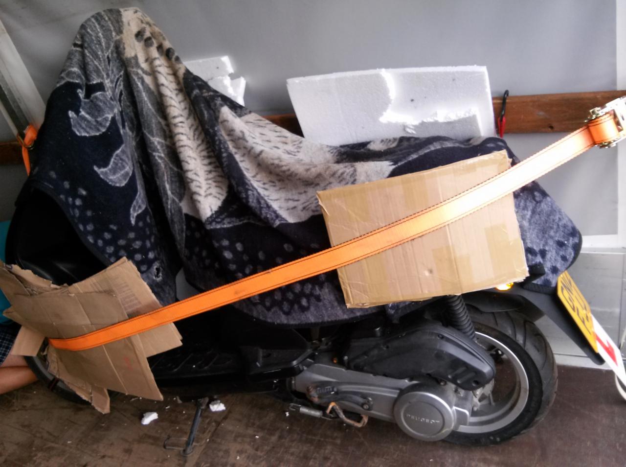 Mopeda zabezpieczono do transportu: koc, kartony, pas transportowy. Widoczny fragment podłogi i ściana. Niestandardowe zabezpieczenie ładunku.