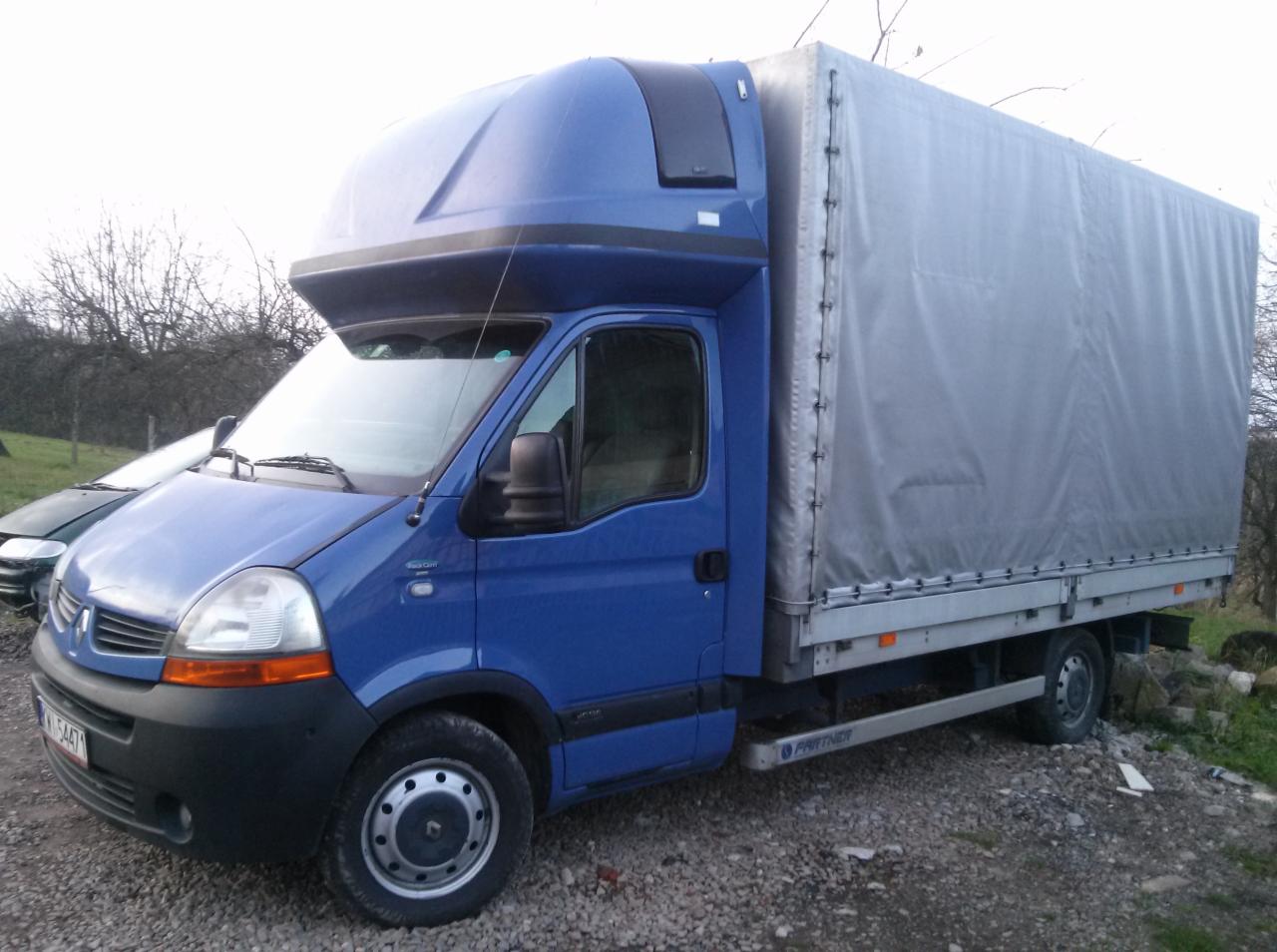 Niebieski bus Renault Master z plandeką na szarej żwirowej nawierzchni, widok z boku. Na pierwszym planie widoczny numer rejestracyjny pojazdu.
