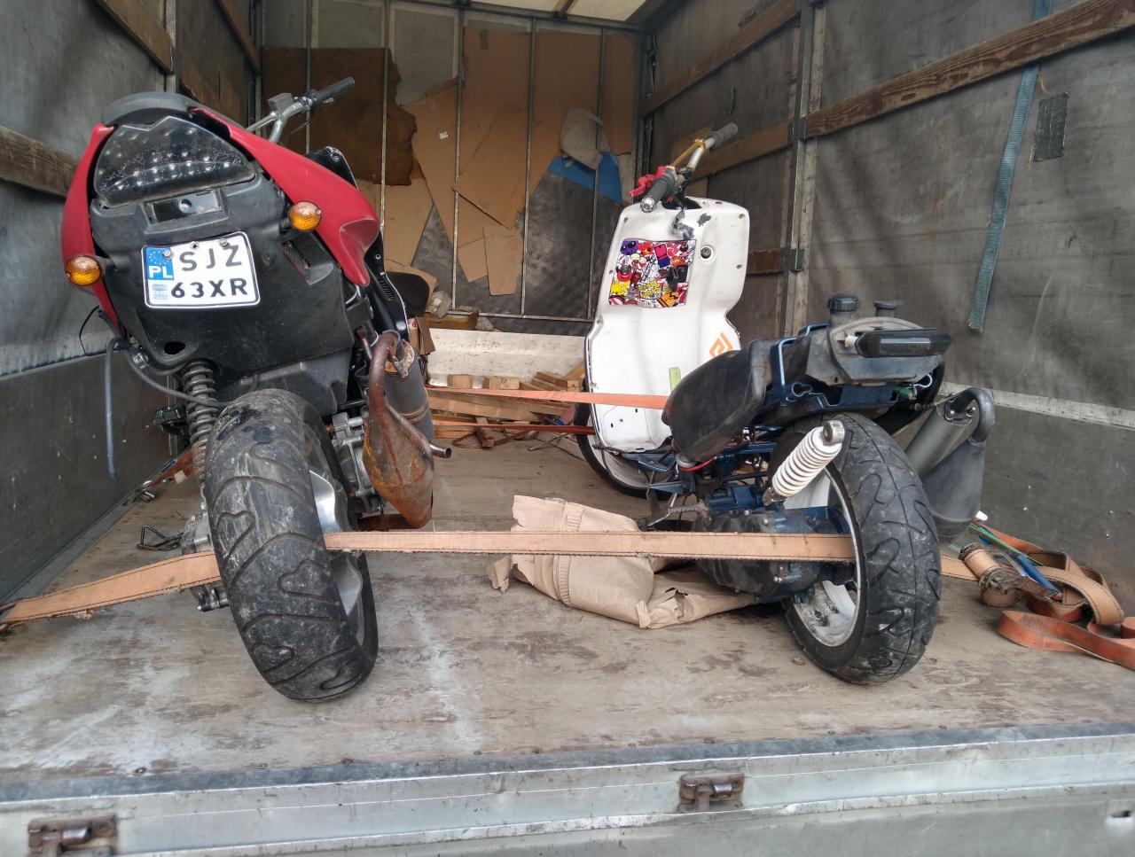 Dwa skutery zabezpieczone pasami transportowymi wewnątrz paki samochodu dostawczego. Jeden skuter z czerwoną obudową i polską tablicą rejestracyjną, drugi biały z naklejkami.