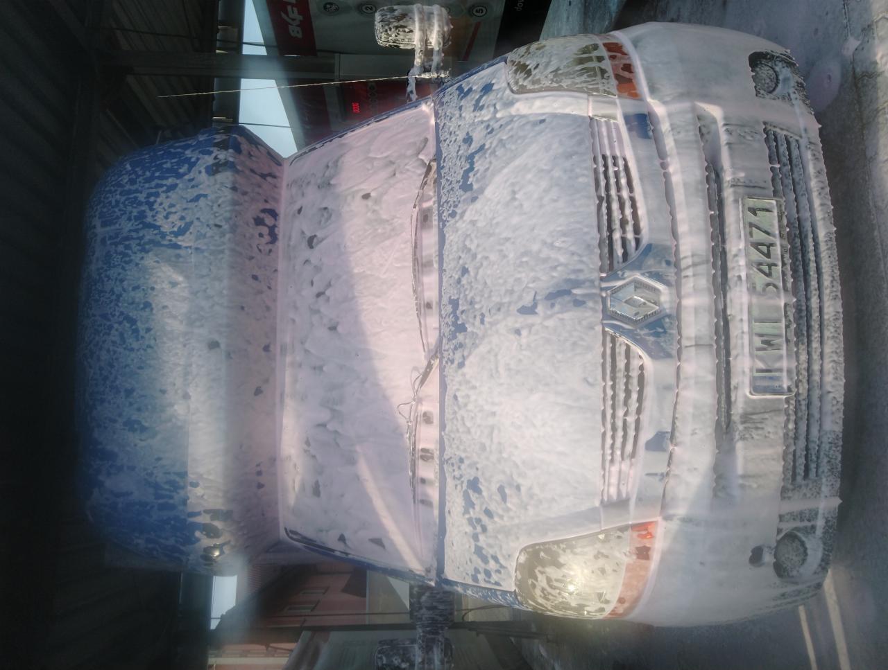 Biały bus pokryty pianą podczas mycia w myjni samochodowej, widoczny fragment tablicy rejestracyjnej.