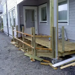 Budowa drewnianego tarasu przy szarym domu z oknami; widoczne elementy konstrukcyjne, podesty i balustrady w trakcie montażu. Na ziemi leżą resztki desek.