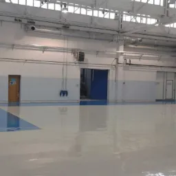 Jasnoszara, błyszcząca posadzka w hali sportowej z niebieskimi akcentami. Widoczne drzwi i instalacje pod sufitem.