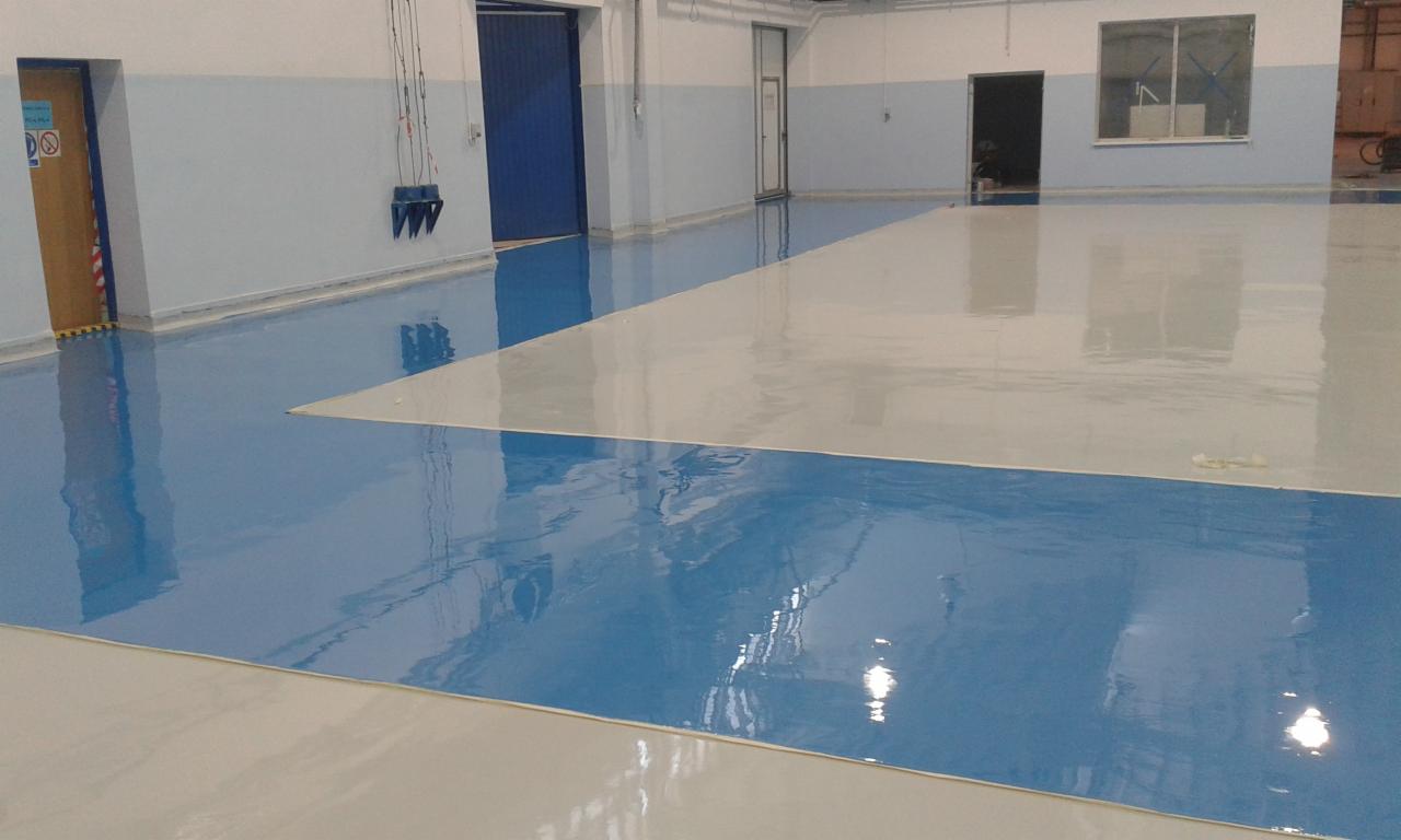 Błękitno-biała, lśniąca posadzka w hali, z widocznymi drzwiami i oknem w tle. Podział kolorystyczny w geometryczne wzory, odbicia światła podkreślają gładką powierzchnię.