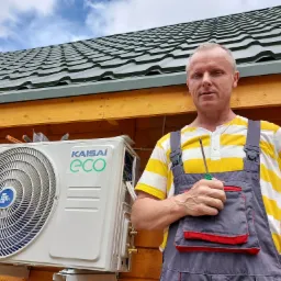 Monter klimatyzacji w pasiastym polo i szarych ogrodniczkach, trzymający śrubokręt, obok zamontowanej jednostki zewnętrznej klimatyzacji Kaisai Eco na drewnianej ścianie budynku z zielonym dachem.