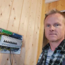 Elektryk w flanelowej koszuli prezentuje zamontowaną skrzynkę bezpiecznikową na ścianie z boazerii, widoczne przewody elektryczne.