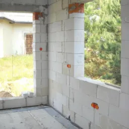 Surowy stan budynku w trakcie budowy, widoczne białe bloczki, okno z widokiem na zieleń, pomarańczowe puszki pod gniazdka i kable w czarnych peszlach ułożone wzdłuż ściany.