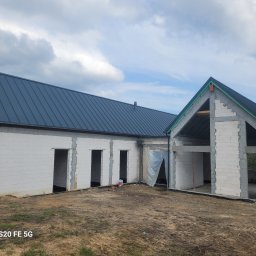 Firma Ogólnobudowlana Mateusz Korbanek - Surowy, biały dom z dachem z blachy trapezowej w trakcie budowy. Widoczne otwory okienne i drzwiowe, teren wokół w stanie naturalnym, pochmurne niebo w tle.