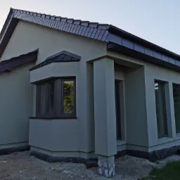 Nowoczesny dom z szarą elewacją i ciemną dachówką, widok z zewnątrz, okna z ciemnymi ramami, teren wokół domu w trakcie prac wykończeniowych.