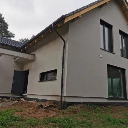 Nowoczesny dom jednorodzinny w trakcie wykończenia elewacji, z widocznym orynnowaniem i stolarką okienną w kolorze antracytowym, na tle pochmurnego nieba.