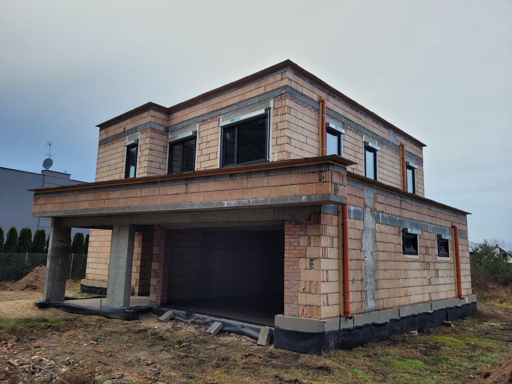 Dwukondygnacyjny dom w trakcie budowy, widoczne mury z pustaków ceramicznych, okna wstawione, brak ocieplenia, rury spustowe.