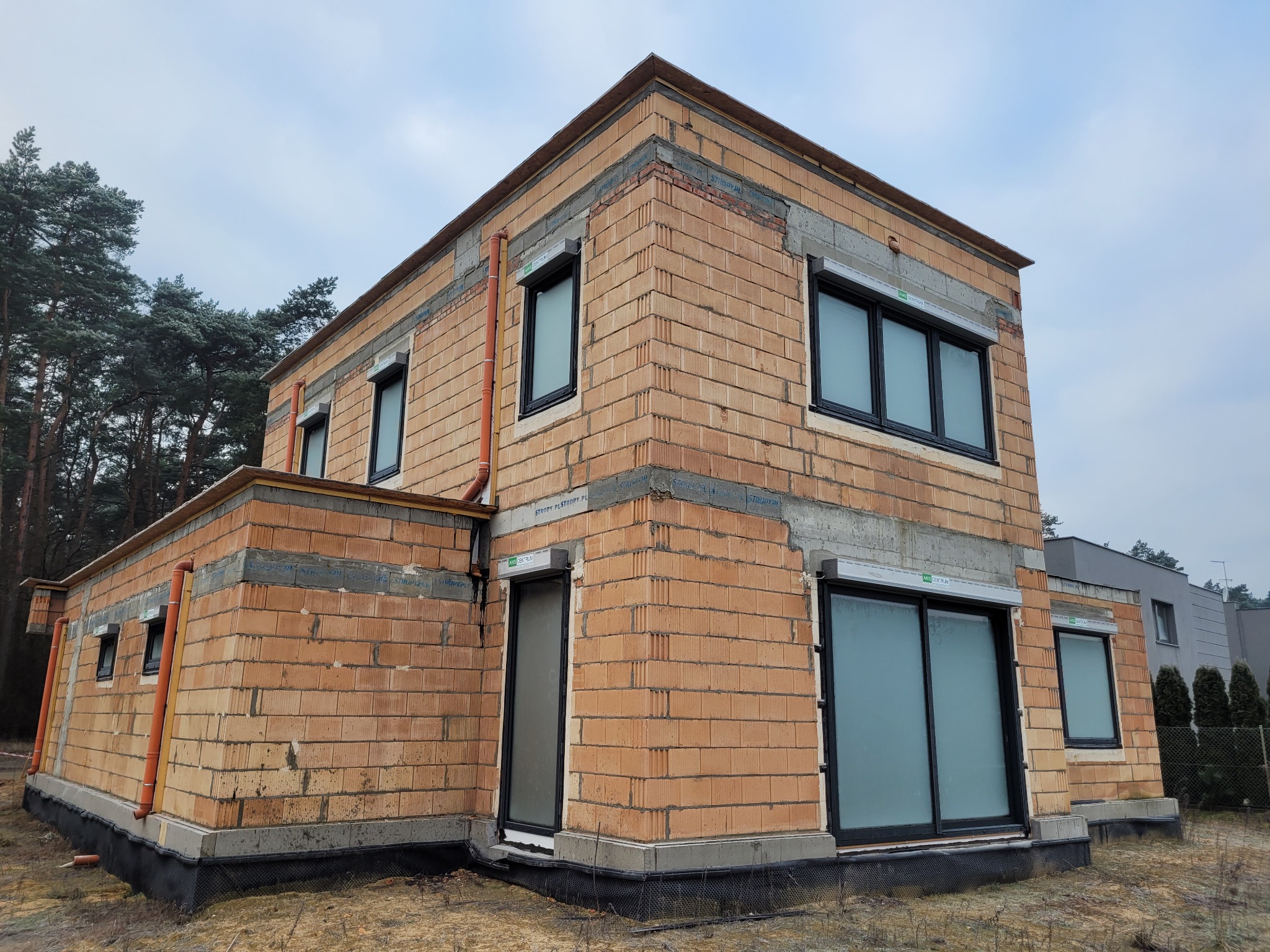 Dwukondygnacyjny dom w trakcie budowy, ściany z czerwonej cegły kratówki, czarne okna z roletami, widoczne przewody wentylacyjne. Stan surowy otwarty, tło: drzewa i pochmurne niebo.