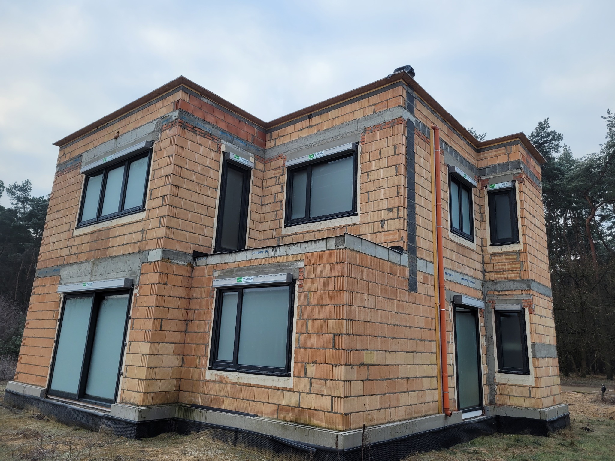Dwukondygnacyjny, surowy dom z cegły ceramicznej z zamontowanymi oknami w czarnych ramach. Widoczne elementy szalunków i zbrojenia. Budowa w toku, stan surowy otwarty.