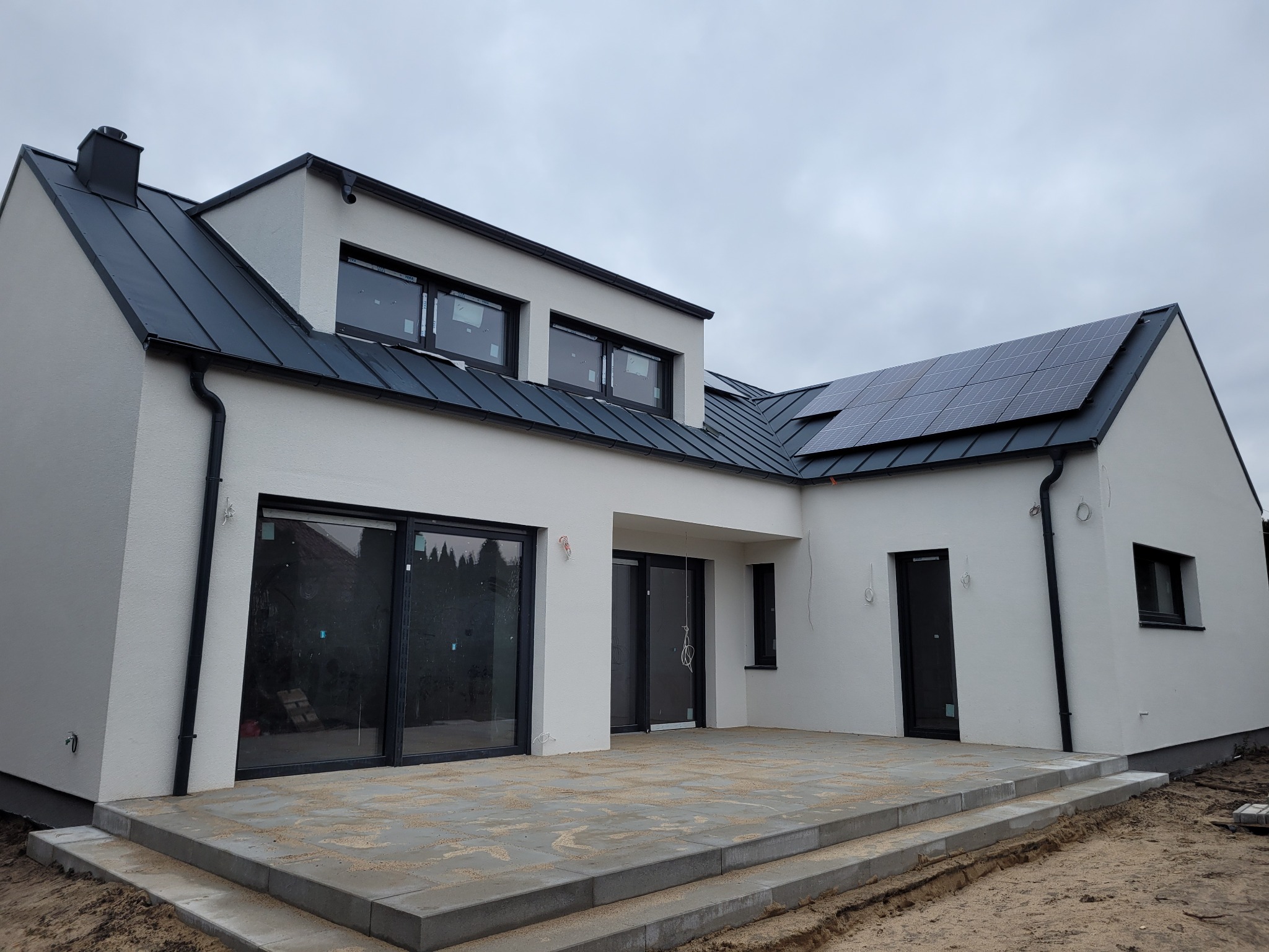 Nowoczesny dom z białymi ścianami, antracytowym dachem z panelami słonecznymi i dużymi oknami, widok na taras z kostki brukowej w pochmurny dzień.