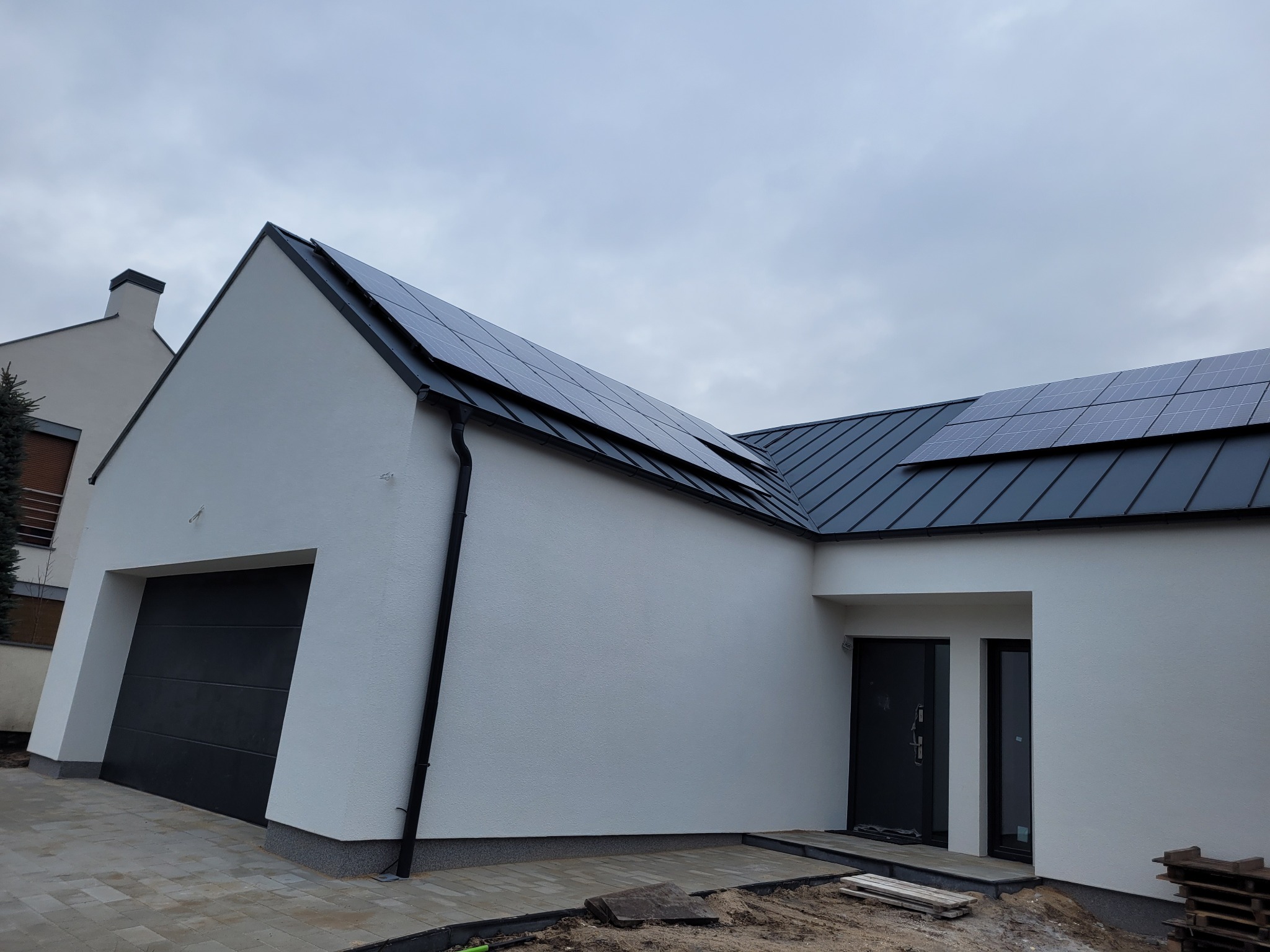 Nowoczesny dom z panelami słonecznymi na dachu i czarną bramą garażową. Elewacja w jasnym odcieniu, widoczne elementy wykończenia terenu. Pochmurne niebo w tle.