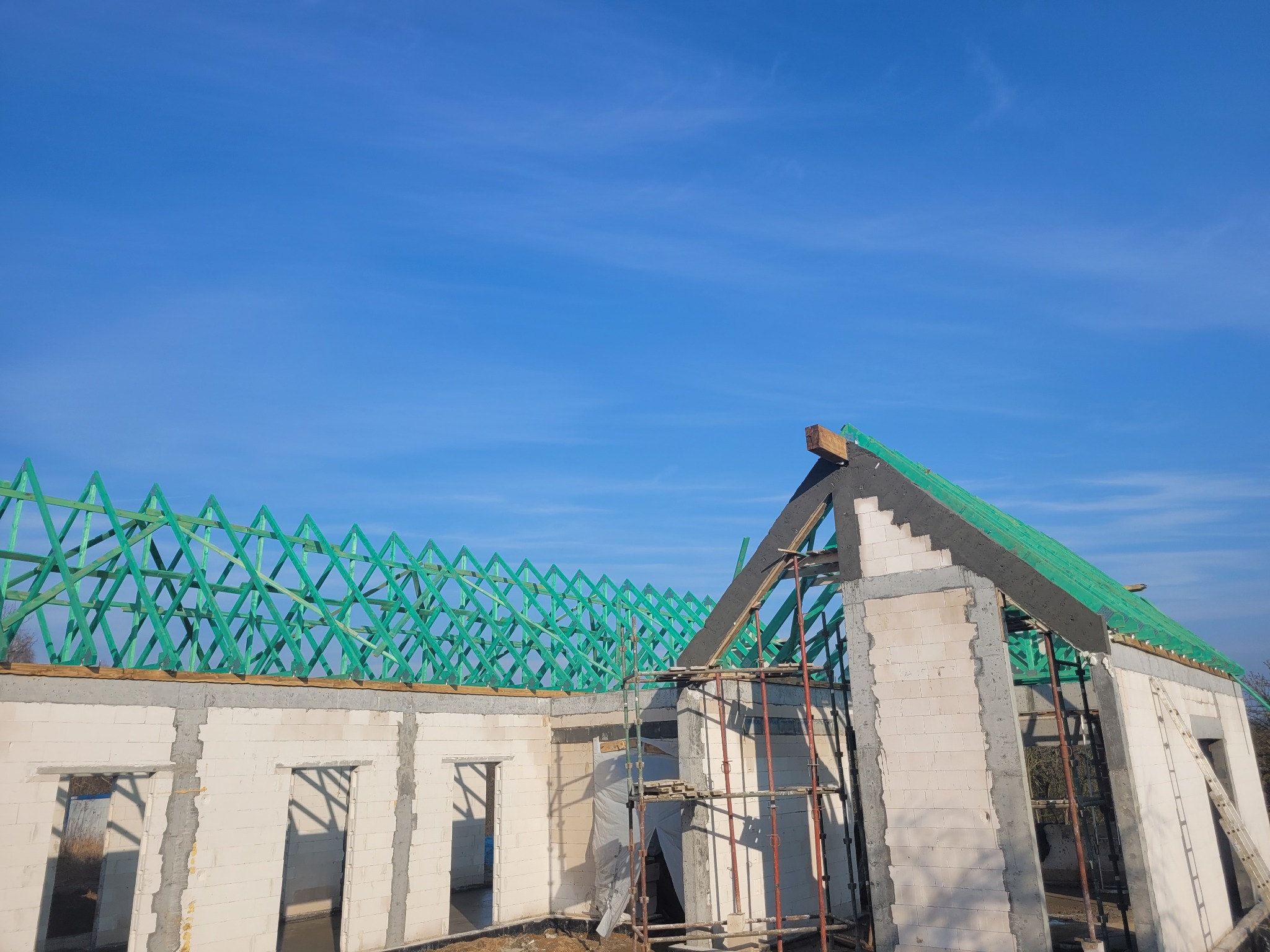 Wznoszenie domu z białych bloczków: ściany parteru, zielona więźba dachowa, rusztowania. Widok na budowę w słoneczny dzień, błękitne niebo w tle.